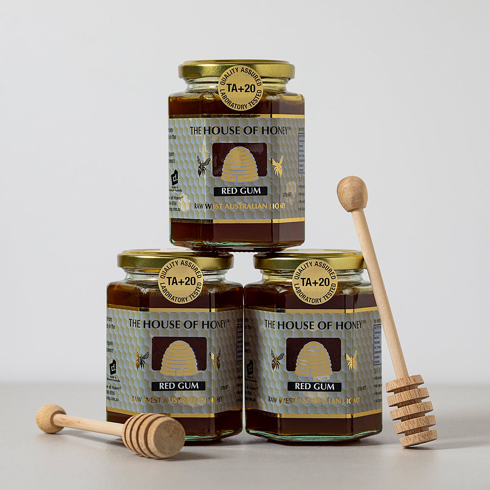 Three jars of 'The House of Honey' red gum honey with honeycombs on labels, accompanied by wooden honey dipper sticks.