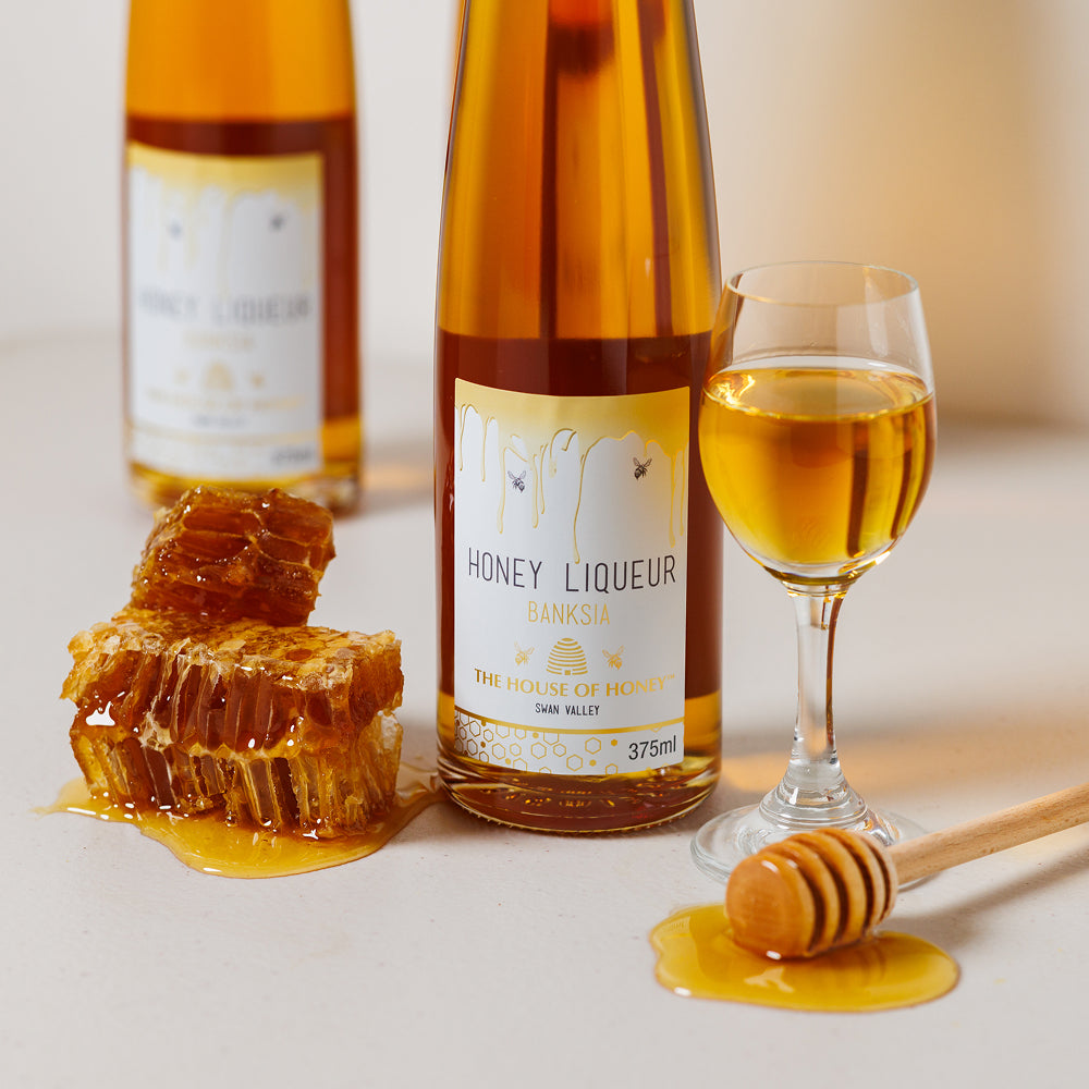 Bottle of honey liqueur with a glass filled with the drink, honeycomb, and dipper on a light surface.