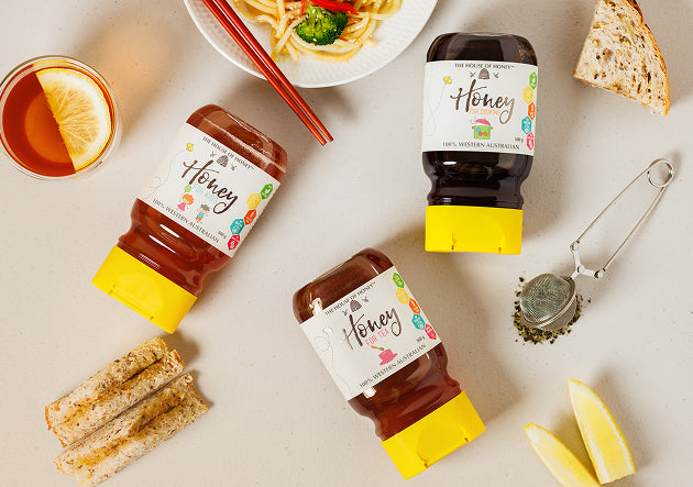 Three bottles of honey with colorful labels on a table with food items.